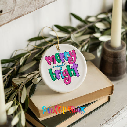 Round ceramic Christmas ornament featuring bright groovy Merry and Bright lettering in pink teal and purple, styled on books with holiday greenery