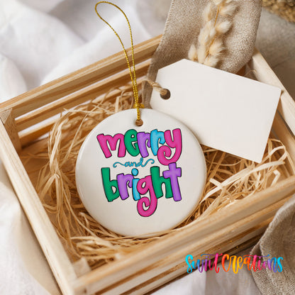 Round ceramic Christmas ornament featuring bright groovy Merry and Bright lettering in pink teal and purple, presented in a rustic wooden gift crate