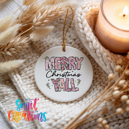 Round ceramic Christmas ornament featuring patterned Merry Christmas Y’all lettering with pink and green accents, styled beside a candle and cozy knit blanket