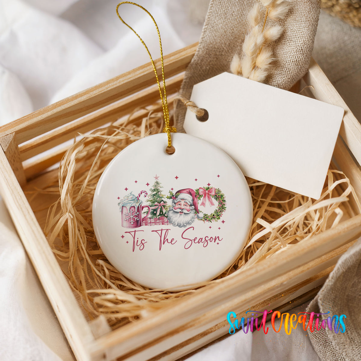Round ceramic Christmas ornament featuring vintage Santa with floral greenery and ’Tis the Season lettering, presented in a rustic wooden gift crate