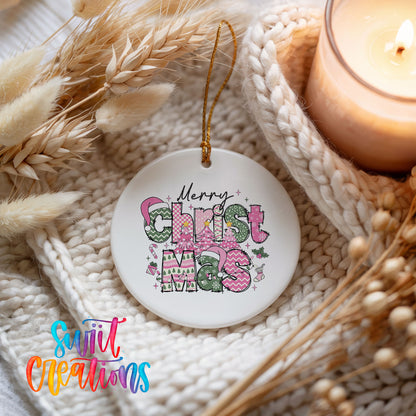 Round ceramic Christmas ornament featuring stacked patterned Merry Christmas lettering in pink green and black, styled beside a candle and cozy knit blanket