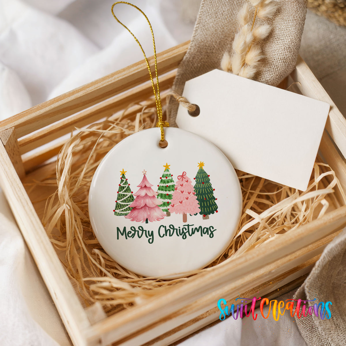 Round ceramic Christmas ornament featuring row of whimsical colorful Christmas trees with Merry Christmas script, presented in a rustic wooden gift crate