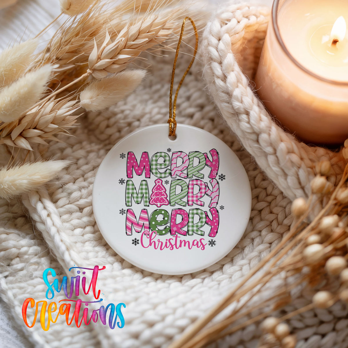 Round ceramic Christmas ornament featuring stacked patterned Merry Merry Merry Christmas lettering in pink and green, styled beside a candle and cozy knit blanket