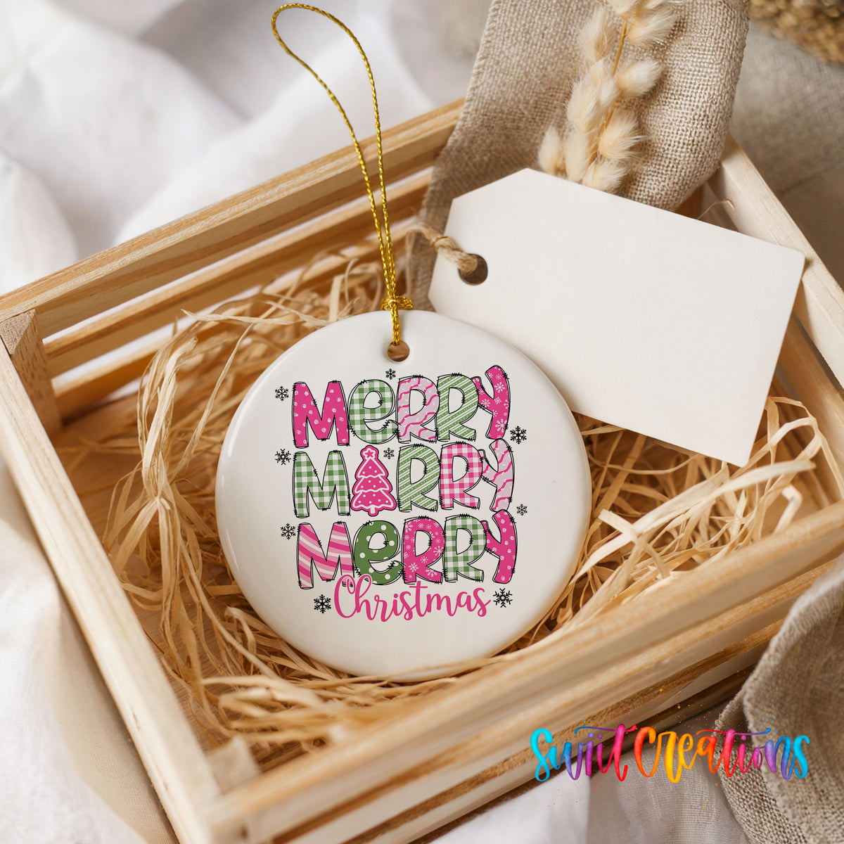 Round ceramic Christmas ornament featuring stacked patterned Merry Merry Merry Christmas lettering in pink and green, presented in a rustic wooden gift crate