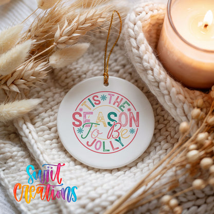 Round ceramic Christmas ornament featuring round pastel ’Tis the Season to Be Jolly holiday typography, styled beside a candle and cozy knit blanket