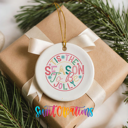 Round ceramic Christmas ornament featuring round pastel ’Tis the Season to Be Jolly holiday typography, placed on a wrapped Christmas gift