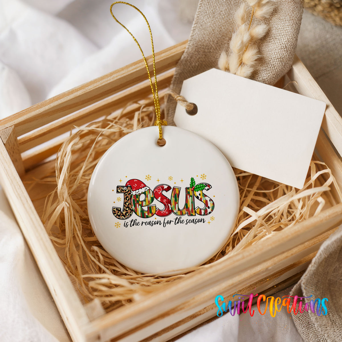 Round ceramic Christmas ornament featuring colorful Jesus Is the Reason for the Season typography, presented in a rustic wooden gift crate