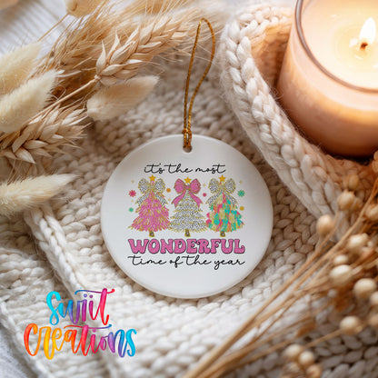 Round ceramic Christmas ornament featuring It’s the Most Wonderful Time of the Year lettering with colorful gifts and trees, styled beside a candle and cozy knit blanket
