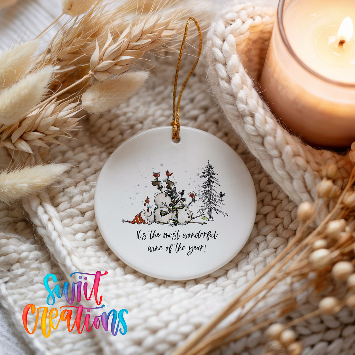 Round ceramic Christmas ornament featuring tipsy snowmen with wine glasses and It’s the Most Wonderful Wine of the Year lettering, styled beside a candle and cozy knit blanket