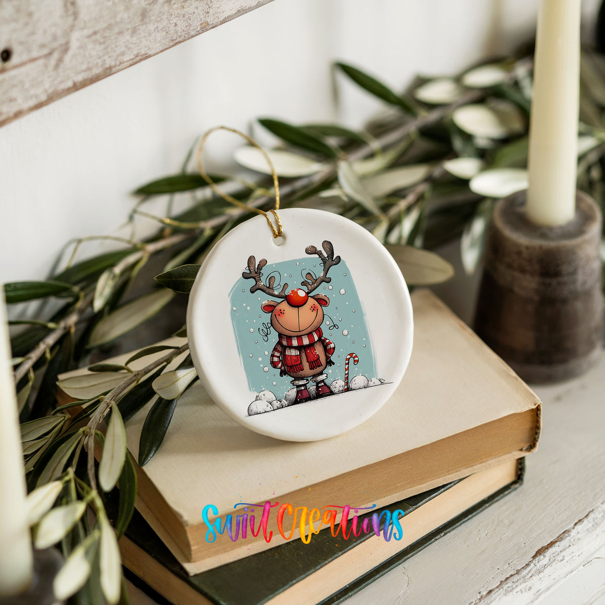 Round ceramic Christmas ornament featuring smiling reindeer in red plaid winter clothes against a snowy blue background, styled on books with holiday greenery