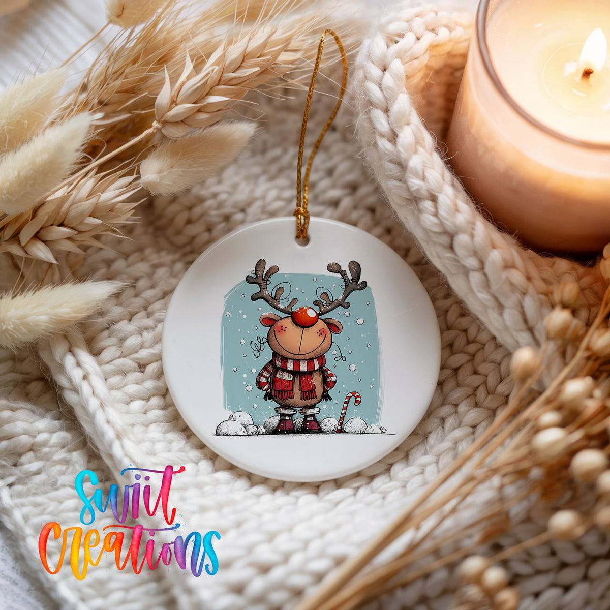 Round ceramic Christmas ornament featuring smiling reindeer in red plaid winter clothes against a snowy blue background, styled beside a candle and cozy knit blanket