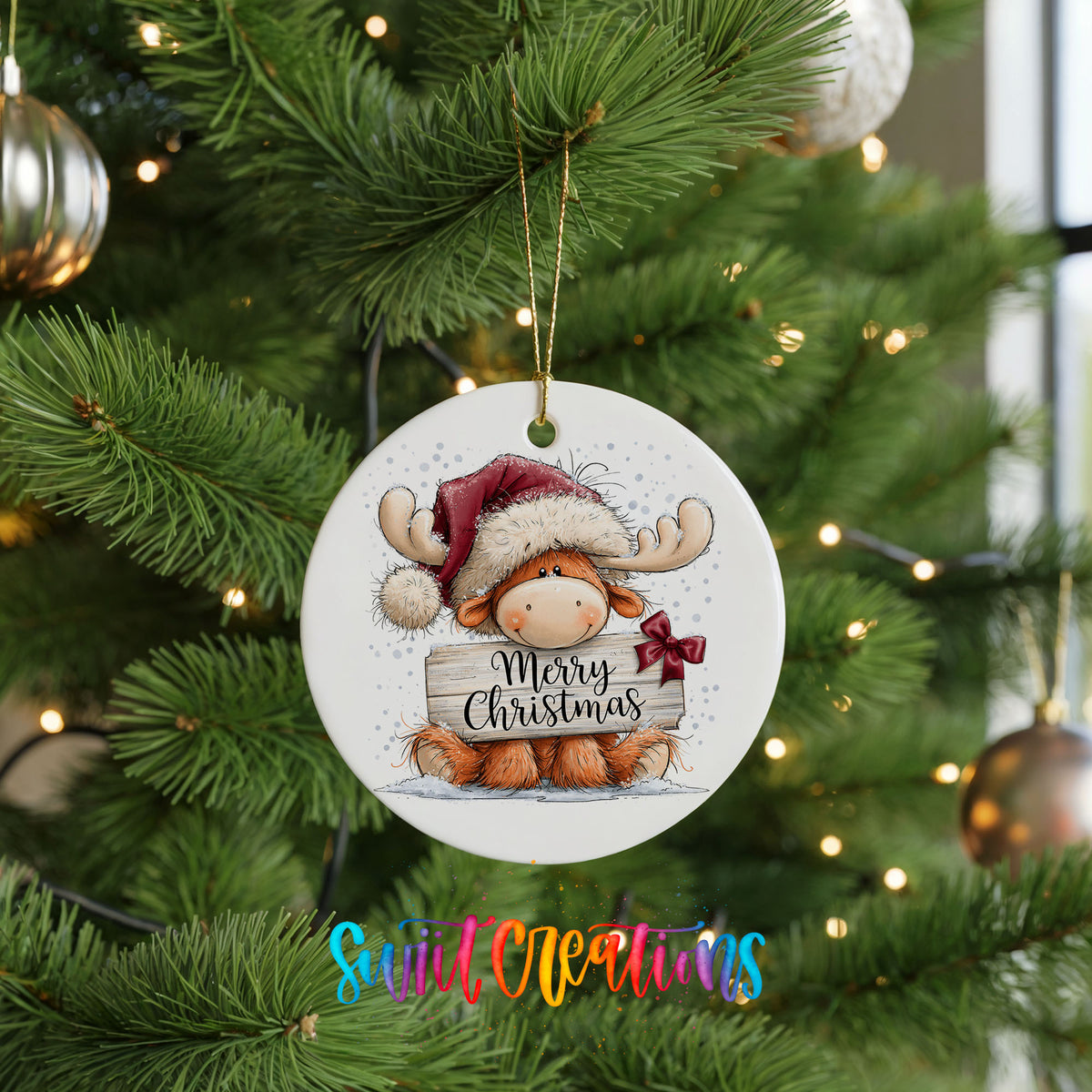 moose in a Santa hat holding a Merry Christmas sign ceramic Christmas ornament hanging on a lit Christmas tree