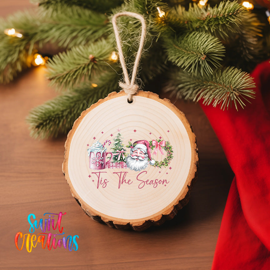 A wooden ornament with a Santa Claus design hangs on a Christmas tree, surrounded by pine branches and twinkling lights.