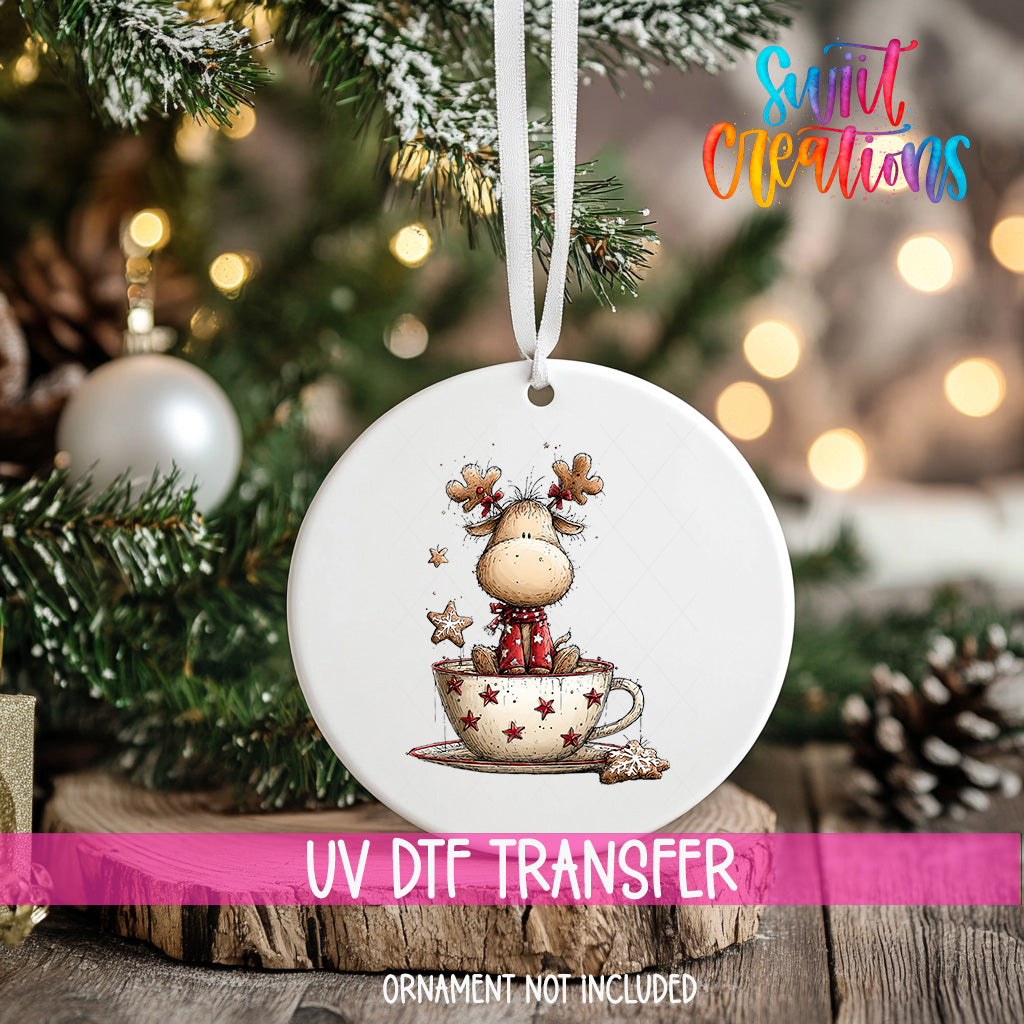 A white ornament with a cartoon reindeer sitting in a cup of coffee, surrounded by snow and twinkling lights, is hanging on a Christmas tree.