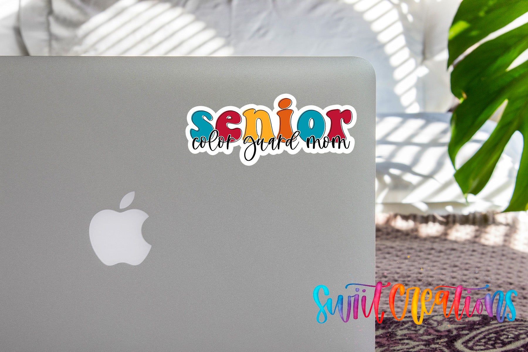 a sticker with the text "senior color guard mom" on the back of a silver laptop, with a plant and a patterned rug in the background.