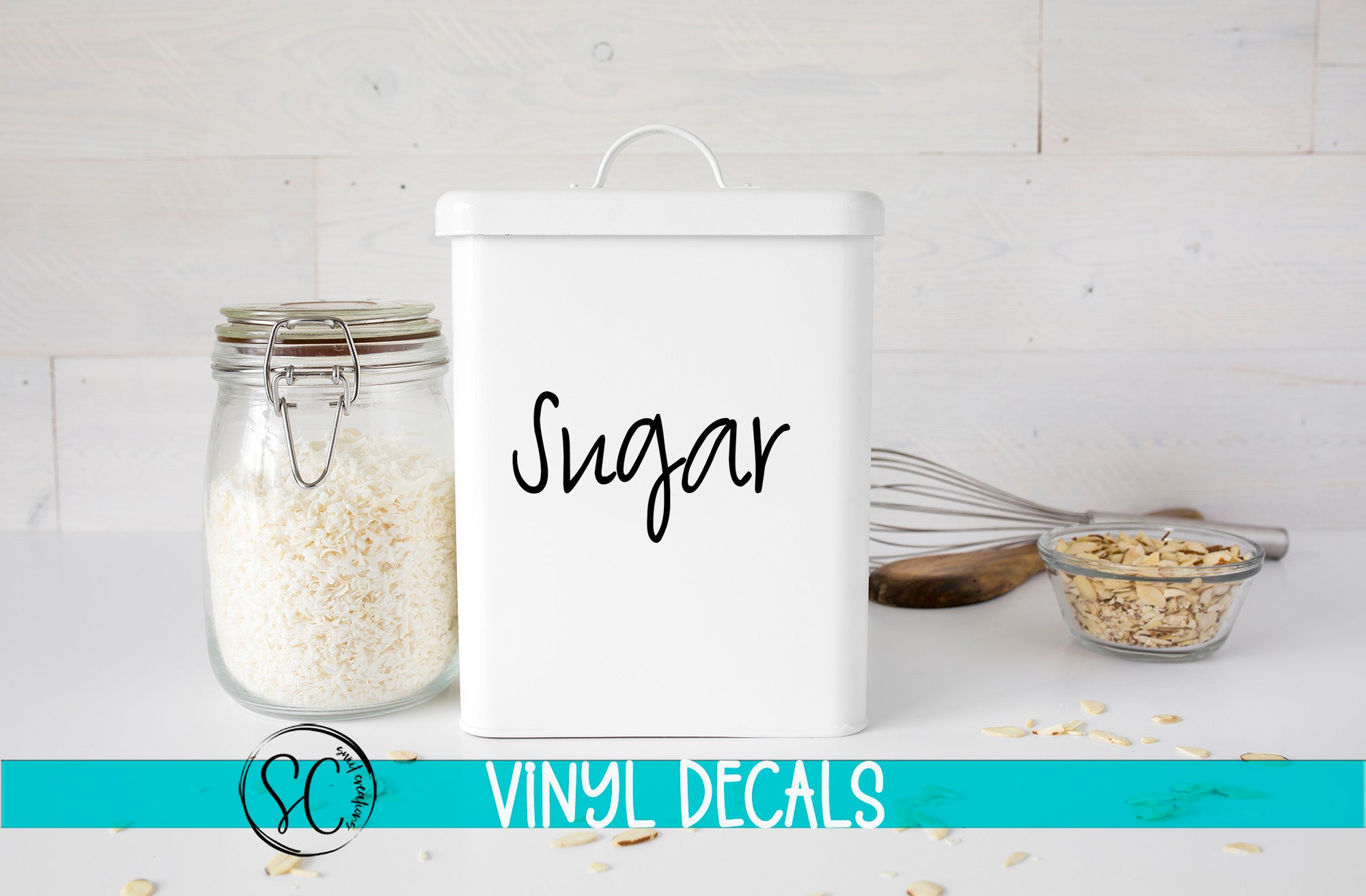 a white canister labeled "Sugar" next to a glass jar filled with white rice, with a whisk and a bowl of nuts on a white countertop.