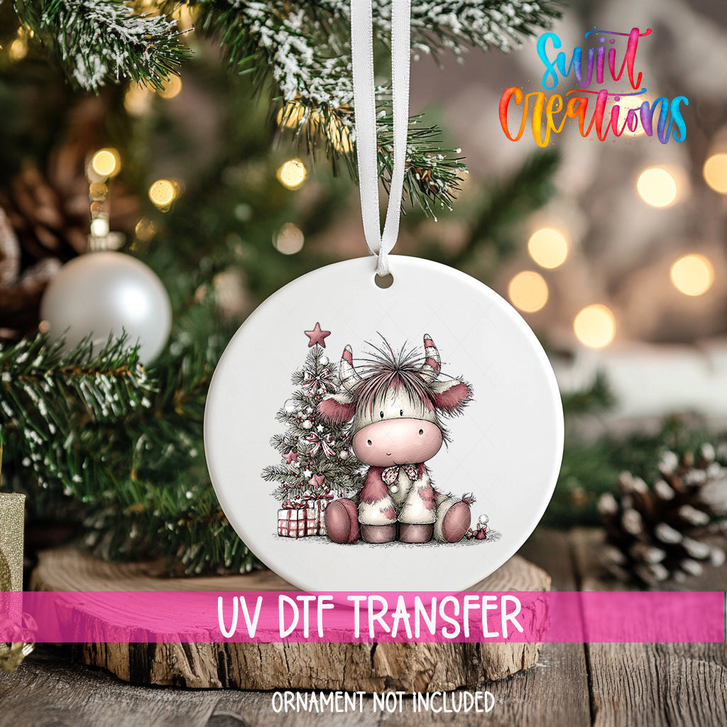 A white ornament with a cow illustration hangs on a Christmas tree, surrounded by pine branches and twinkling lights.
