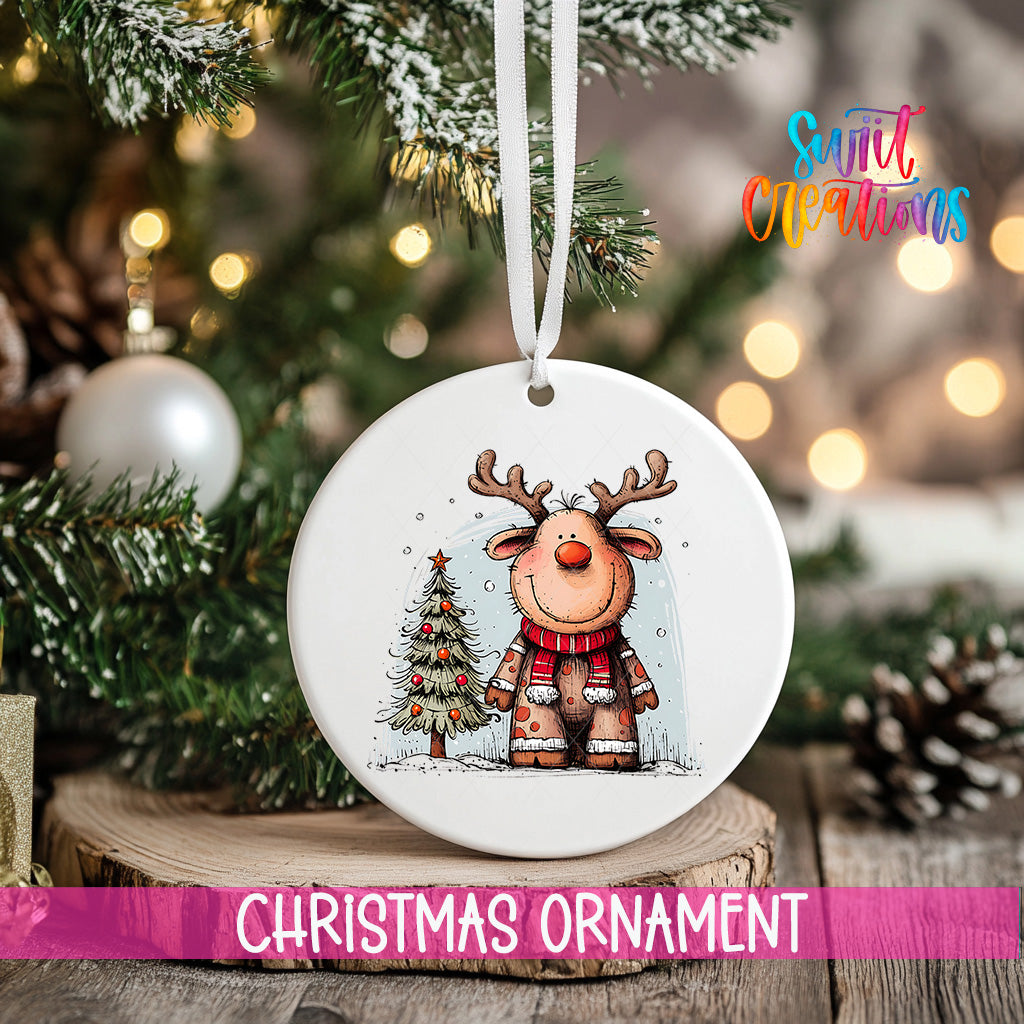 A white Christmas ornament featuring a cartoon reindeer is hanging on a tree branch, with a blurred background of Christmas lights and pine cones.