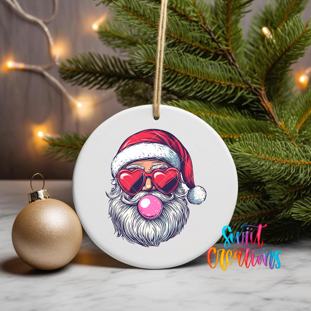 A white ceramic ornament with a Santa Claus face wearing sunglasses and a red hat, along with a pink bubble gum bubble, is displayed on a white surface next to a gold ornament and a Christmas tree.