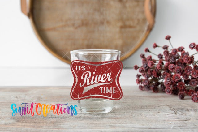 A glass with a red label that says "It's River Time" is placed on a wooden surface, with a bunch of red flowers and a wooden tray in the background.