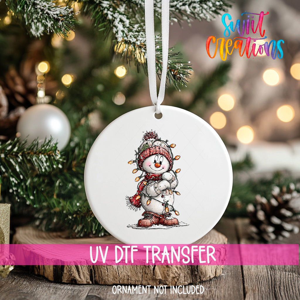 A white ornament with a snowman design is hanging on a Christmas tree, with a pink ribbon around it.
