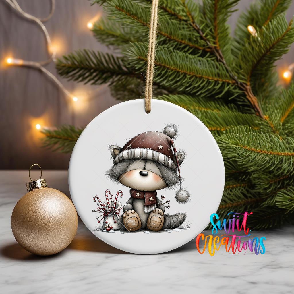 A white ceramic ornament featuring a cartoon raccoon wearing a hat and scarf, sitting on a branch with a Christmas tree in the background.