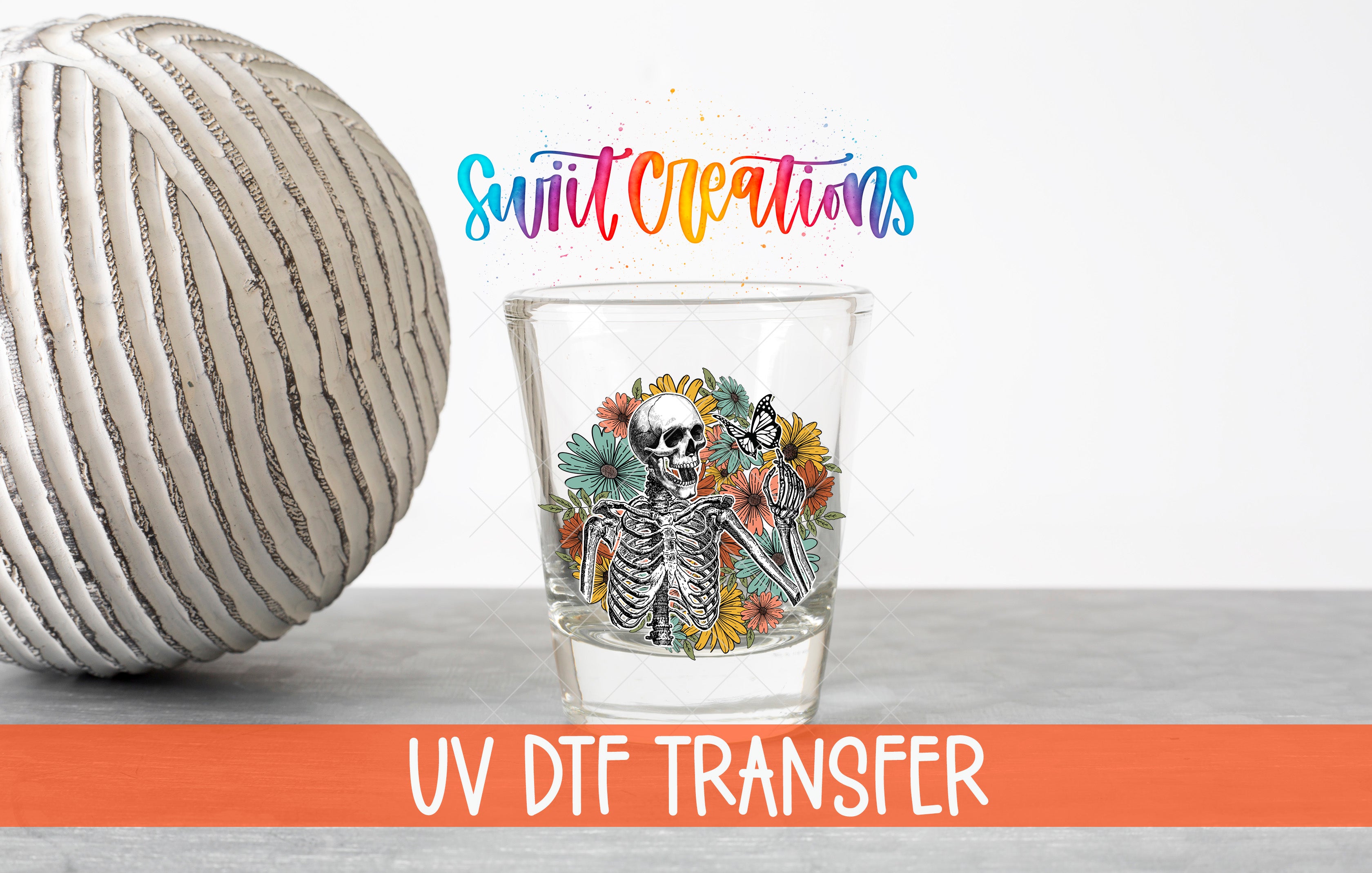 a glass with a skeleton design and flowers, placed next to a white ball with a striped pattern. The text "Swift Creations" is written above the glass, and "UV DFT Transfer" is written below it.