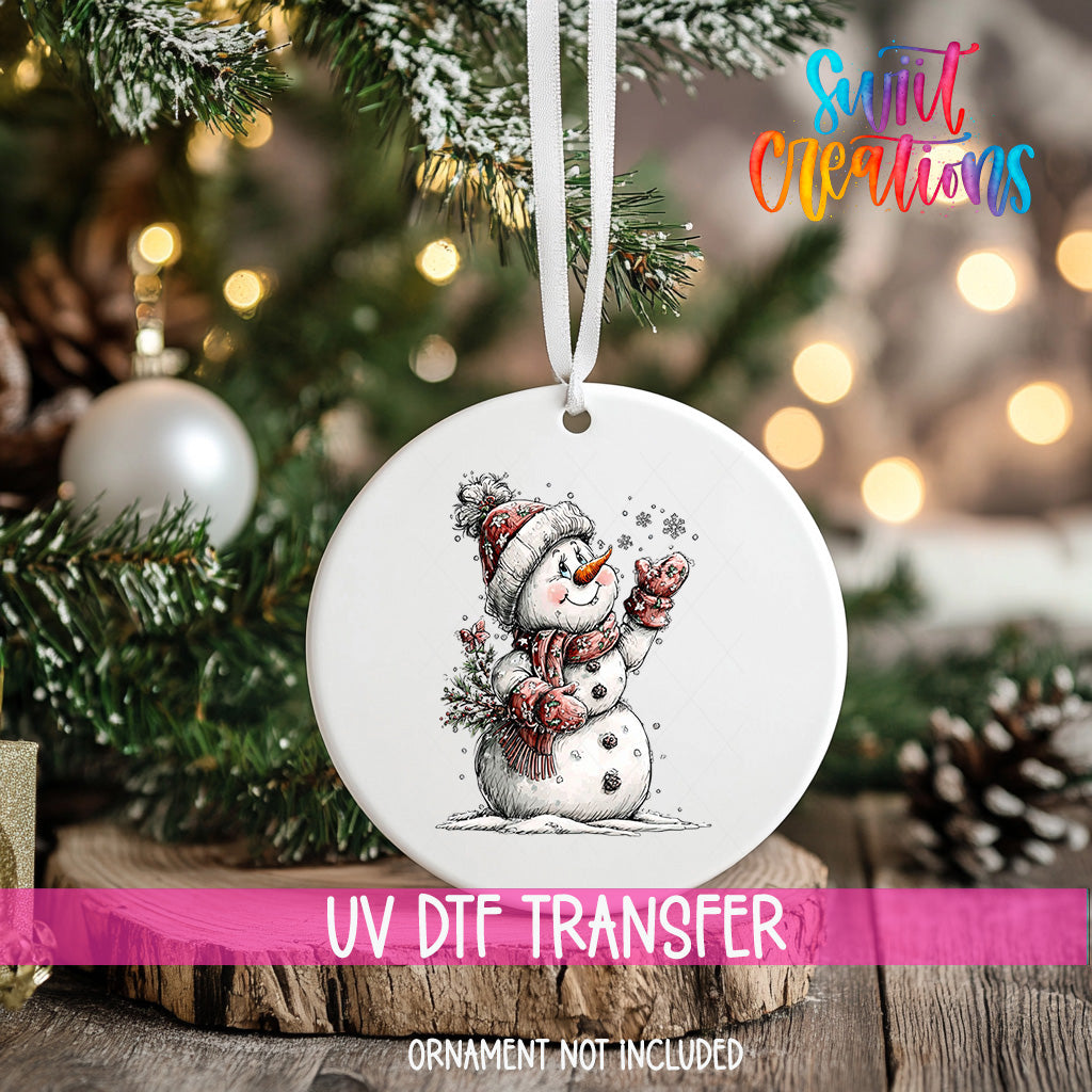 A white ornament with a snowman design hangs on a Christmas tree, surrounded by pine branches and twinkling lights.