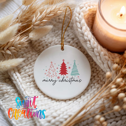 A white ceramic ornament with a Christmas tree design and the text "Merry Christmas" is displayed on a white knitted blanket, with a lit candle and dried flowers in the background.