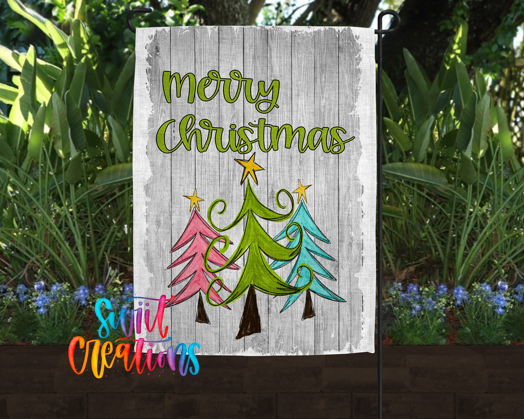 A white wooden sign with a festive Christmas tree design and the text "Merry Christmas" is displayed in front of a garden with green plants and blue flowers.
