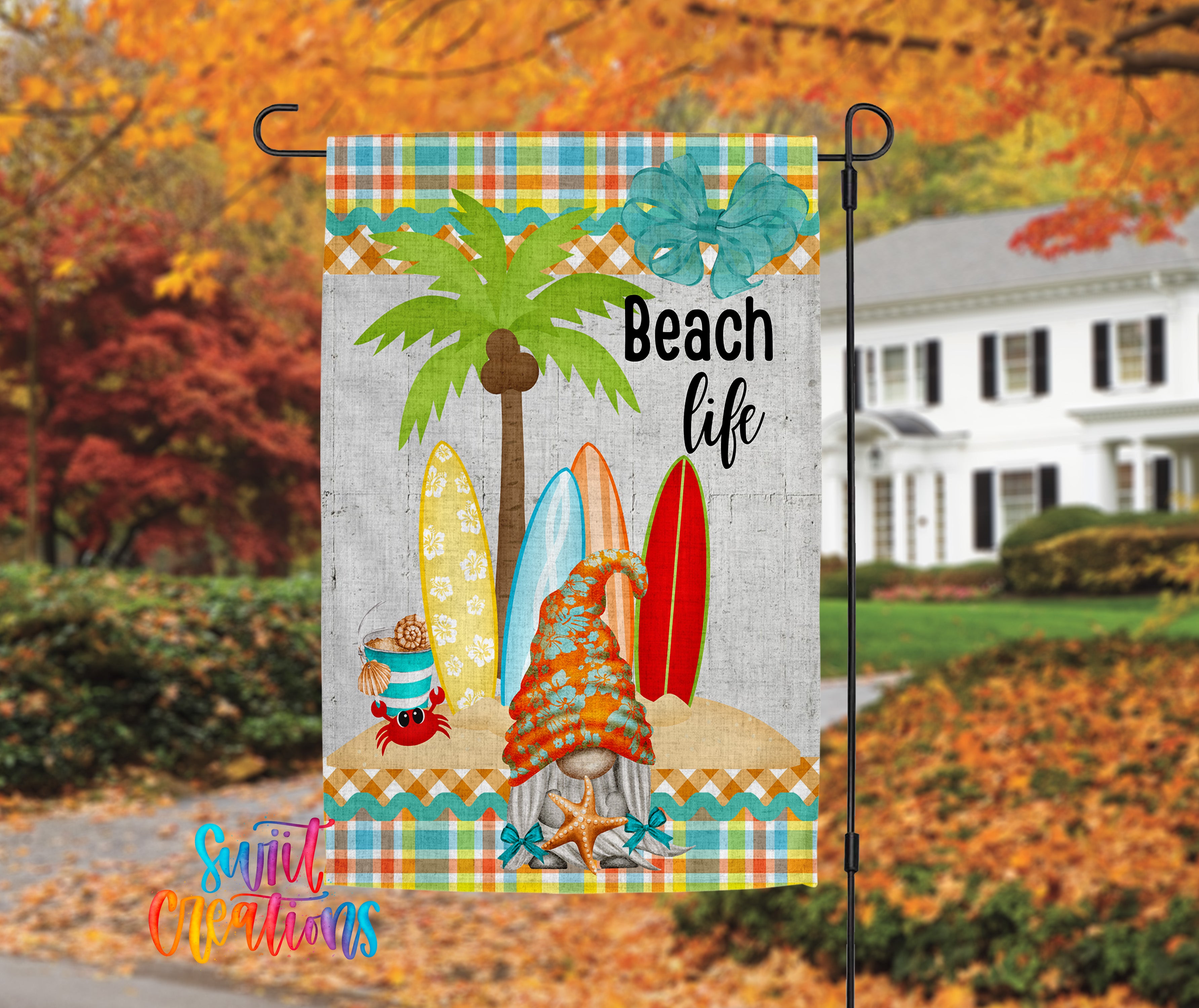 A colorful beach-themed flag with a palm tree, surfboards, and a gnome figure, hanging on a black pole in front of a house with autumn foliage in the background.