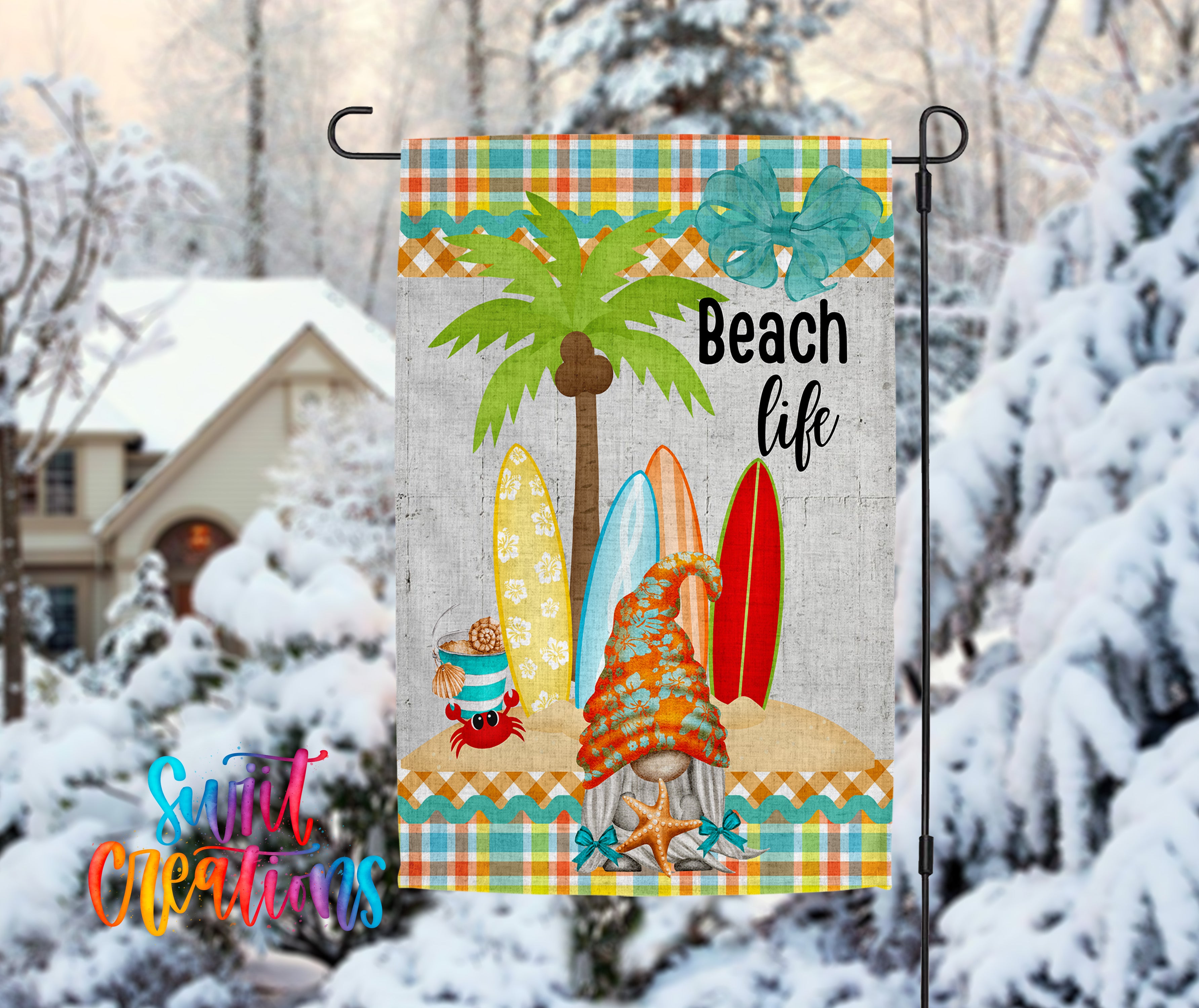 A colorful beach-themed garden flag with a palm tree, surfboards, and a gnome, set against a snowy background with a house and trees.