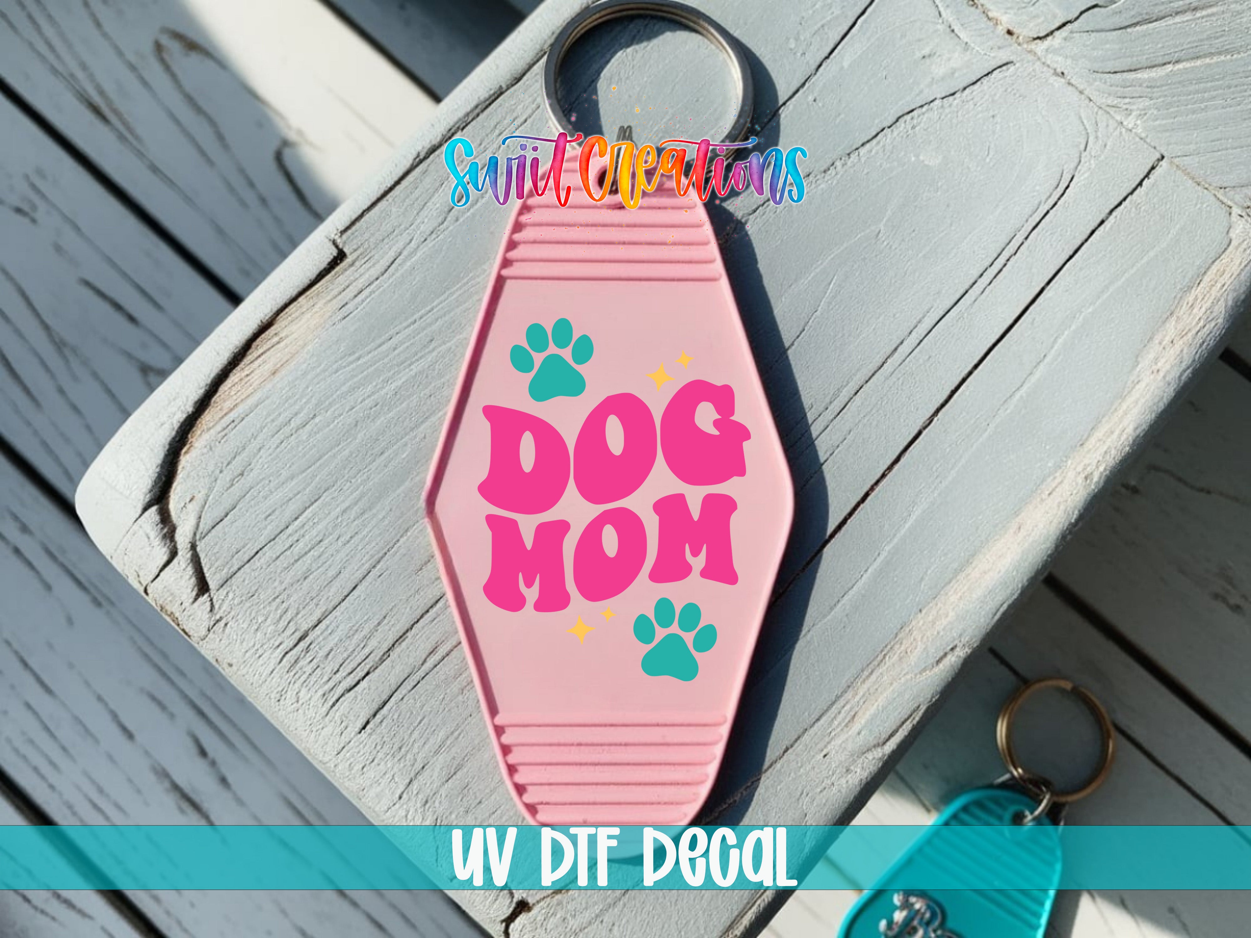 A pink keychain with the text "DOG MOM" printed on it, along with paw prints, is placed on a wooden surface.