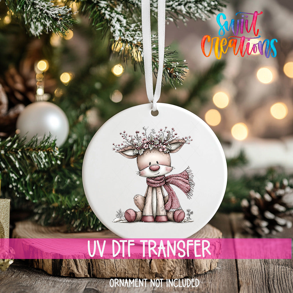 A white ornament with a deer and a scarf is hanging on a Christmas tree, with a pink ribbon around it.