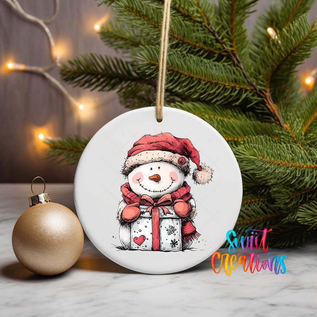A white ceramic ornament featuring a smiling snowman holding a gift box, with a Christmas tree and twinkling lights in the background.
