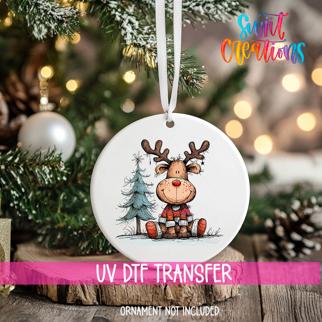 A white ornament with a cartoon reindeer and a Christmas tree is hanging on a tree, with a pink ribbon around it.