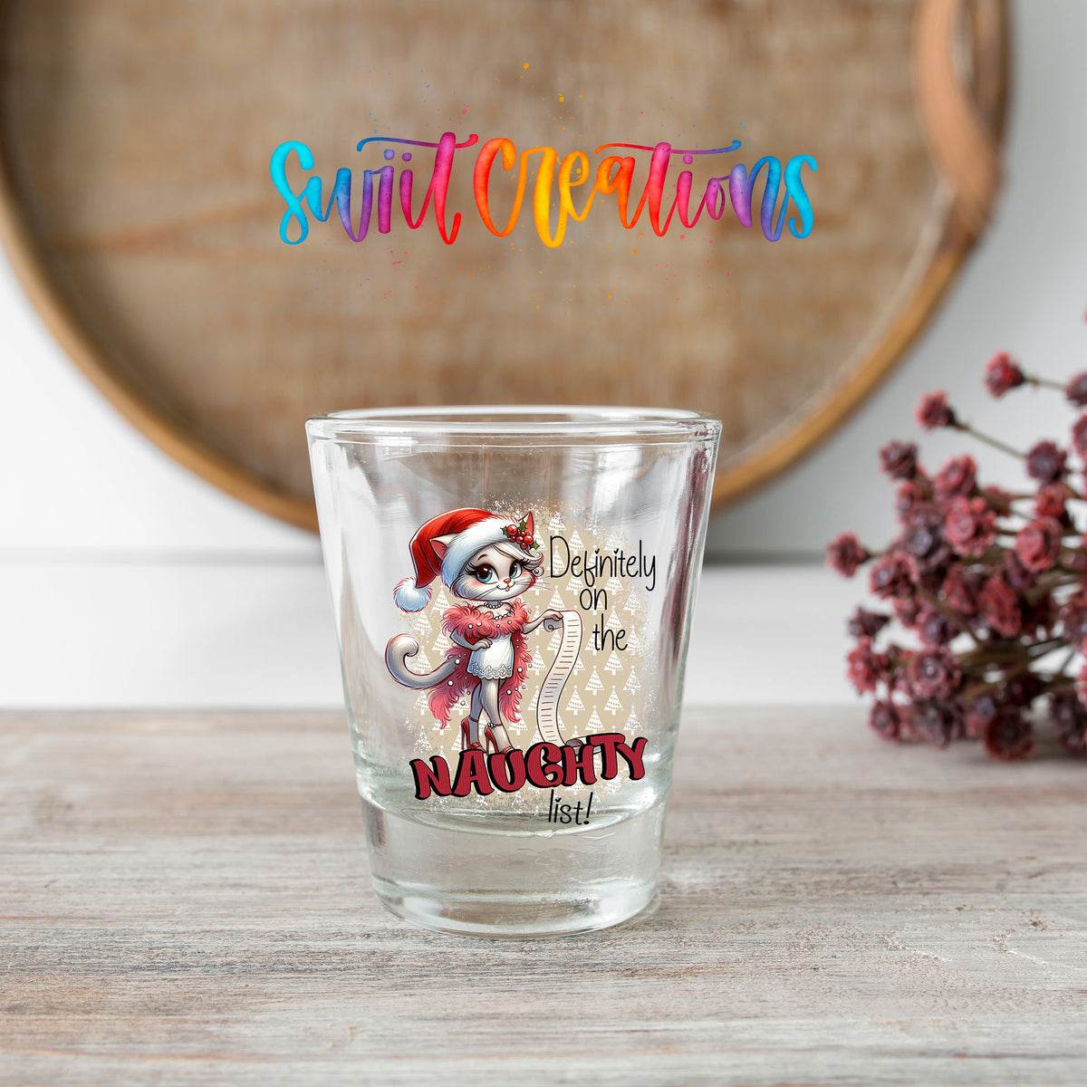 Definitely on the Naughty List Christmas shot glass transfer on a clear glass beside a wooden barrel and dried flowers