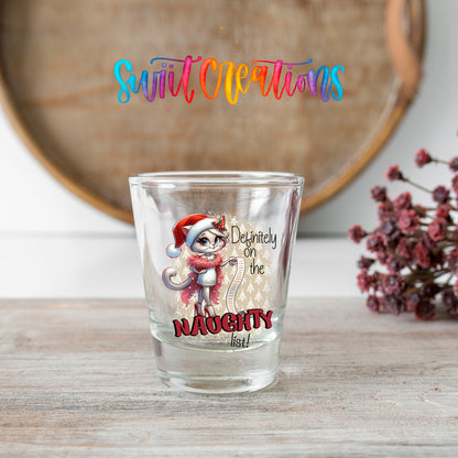 Definitely on the Naughty List Christmas shot glass transfer on a clear glass beside a wooden barrel and dried flowers