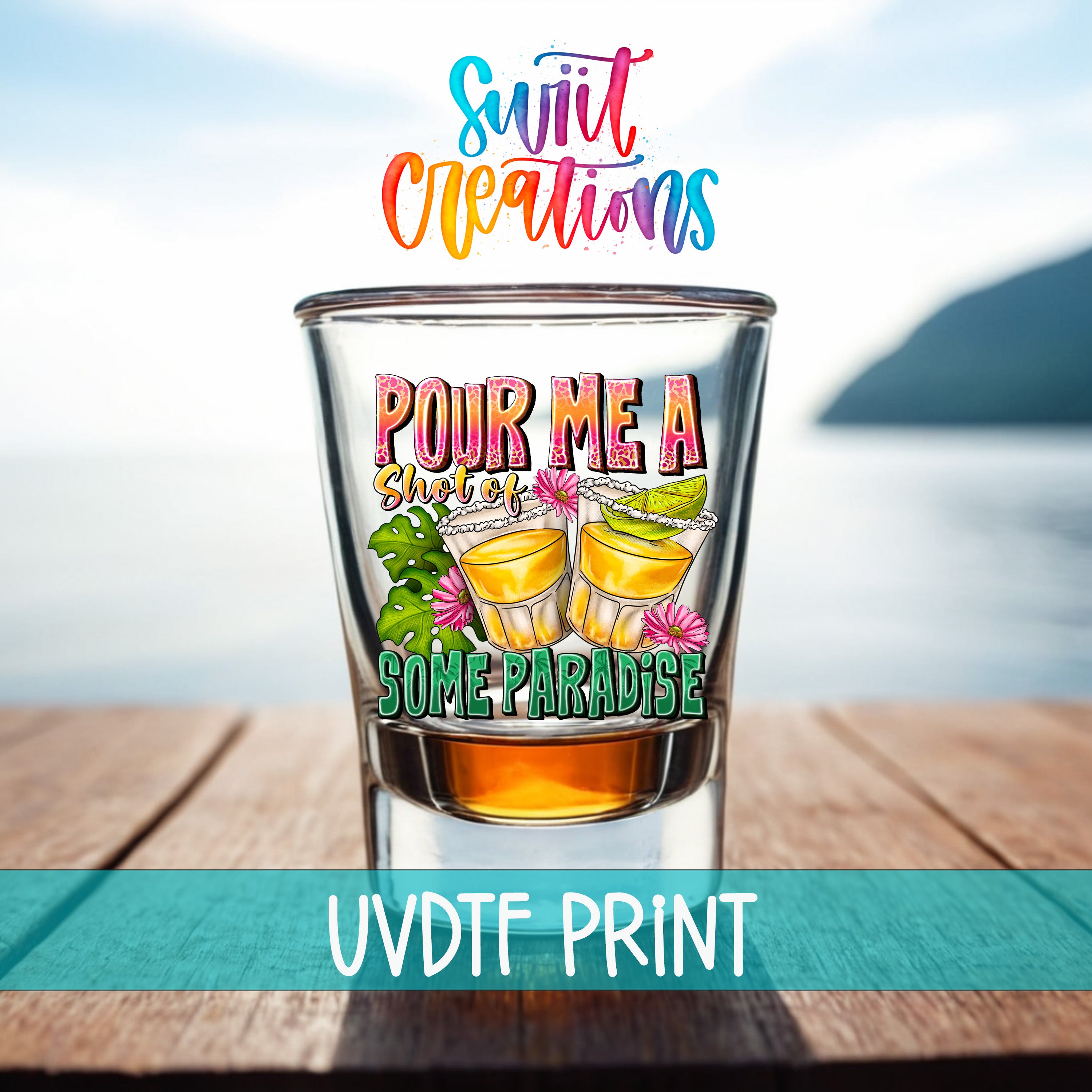 a shot glass with a colorful design and text, placed on a wooden surface with a blurred background that includes a body of water and a mountain.