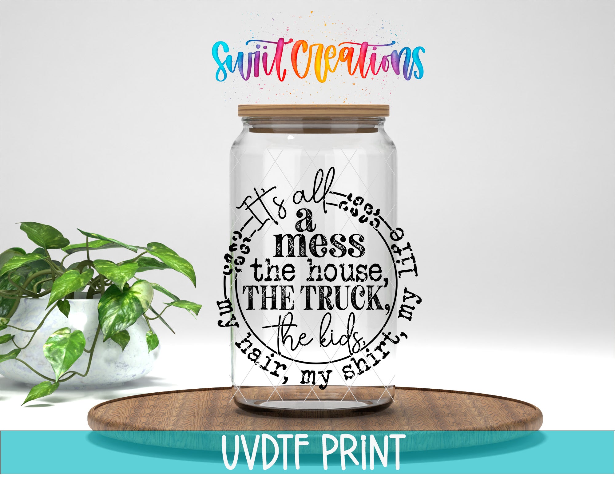 A clear glass jar with a wooden lid, featuring a black and white text design that says "It's all a mess the house the truck the kids". The jar is placed on a wooden tray, and there is a potted plant in the background.