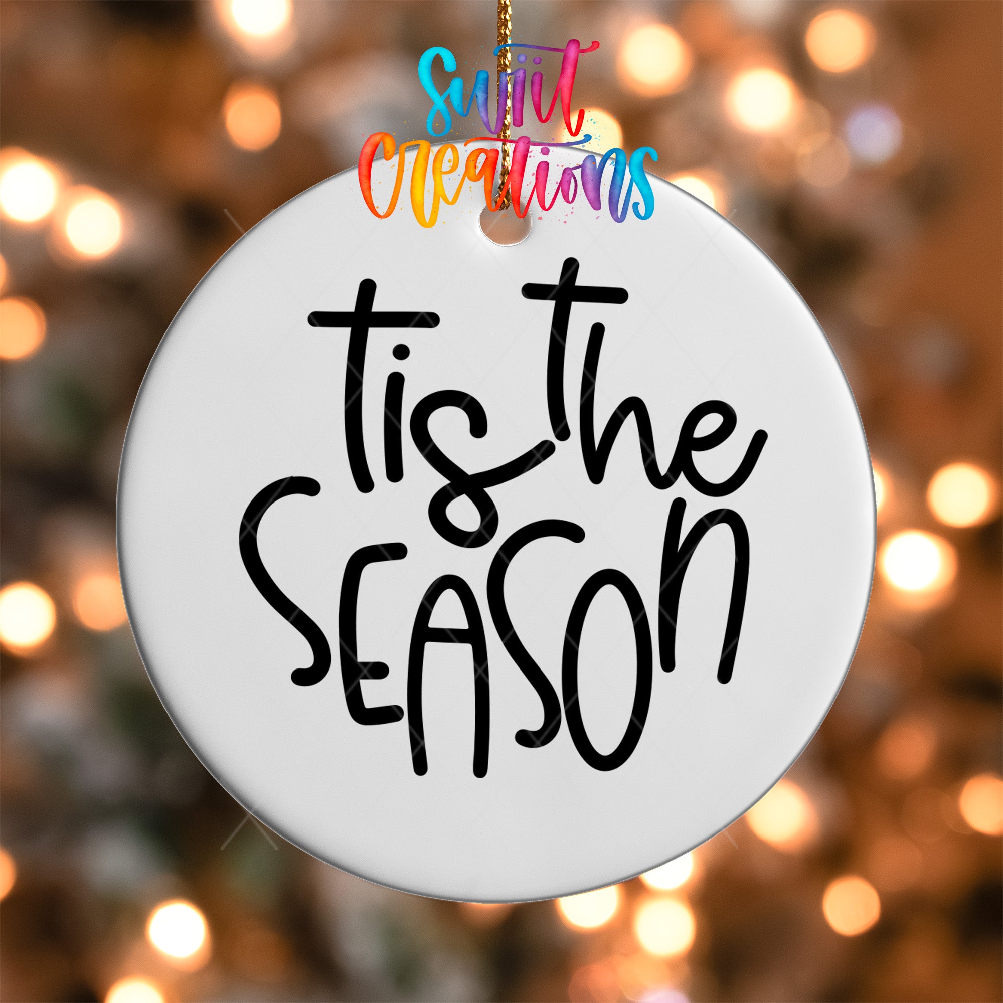 A white circular ornament with the text "Tis the SEASON" written on it, hanging against a background of colorful lights.