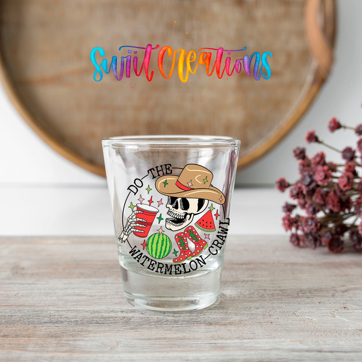 Do The Watermelon Crawl shot glass transfer on a clear glass beside a wooden barrel and dried flowers