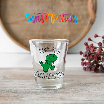 Don't Be a Cuntasaurus shot glass transfer on a clear glass beside a wooden barrel and dried flowers