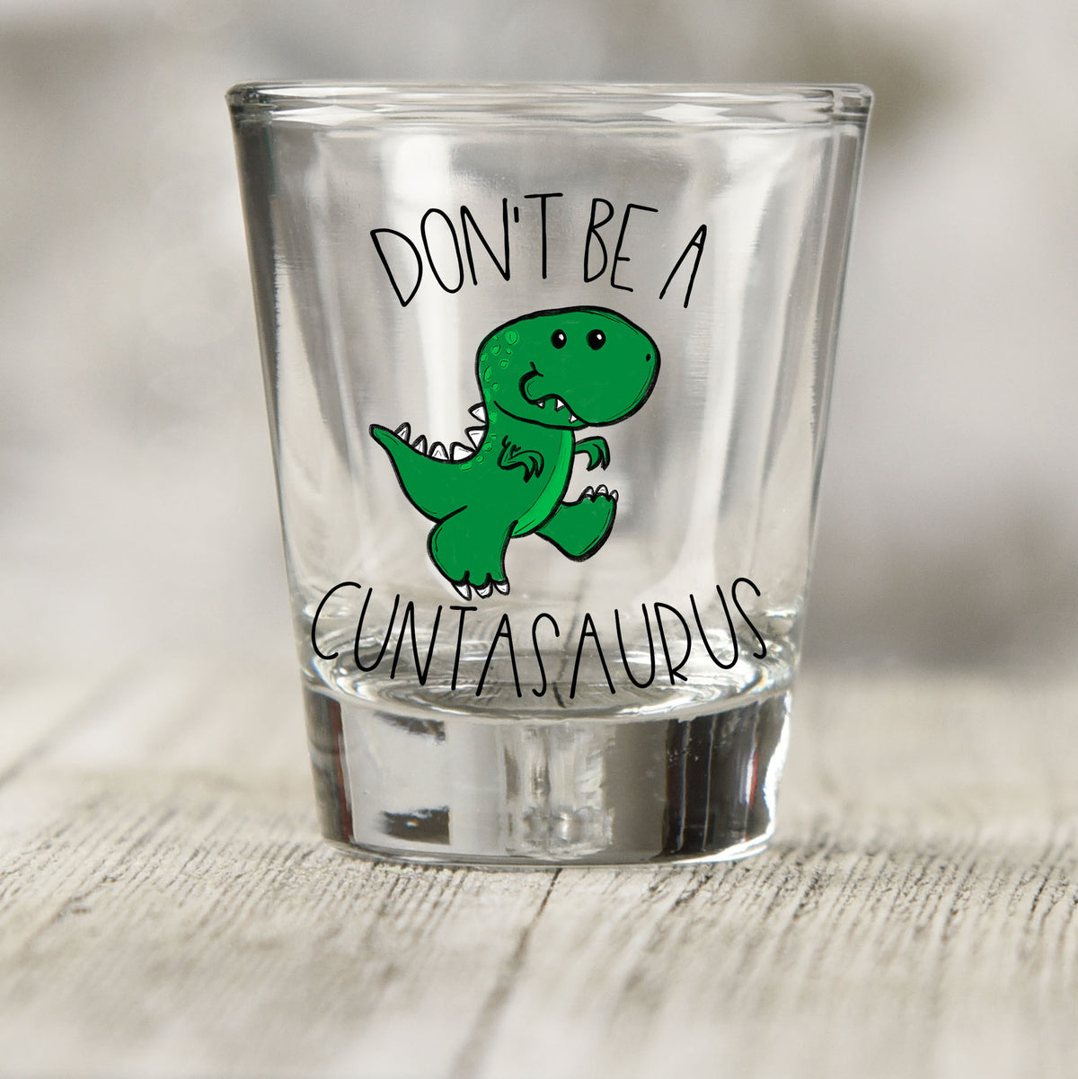 Don't Be a Cuntasaurus shot glass UV DTF transfer with green cartoon dinosaur on a clear shot glass
