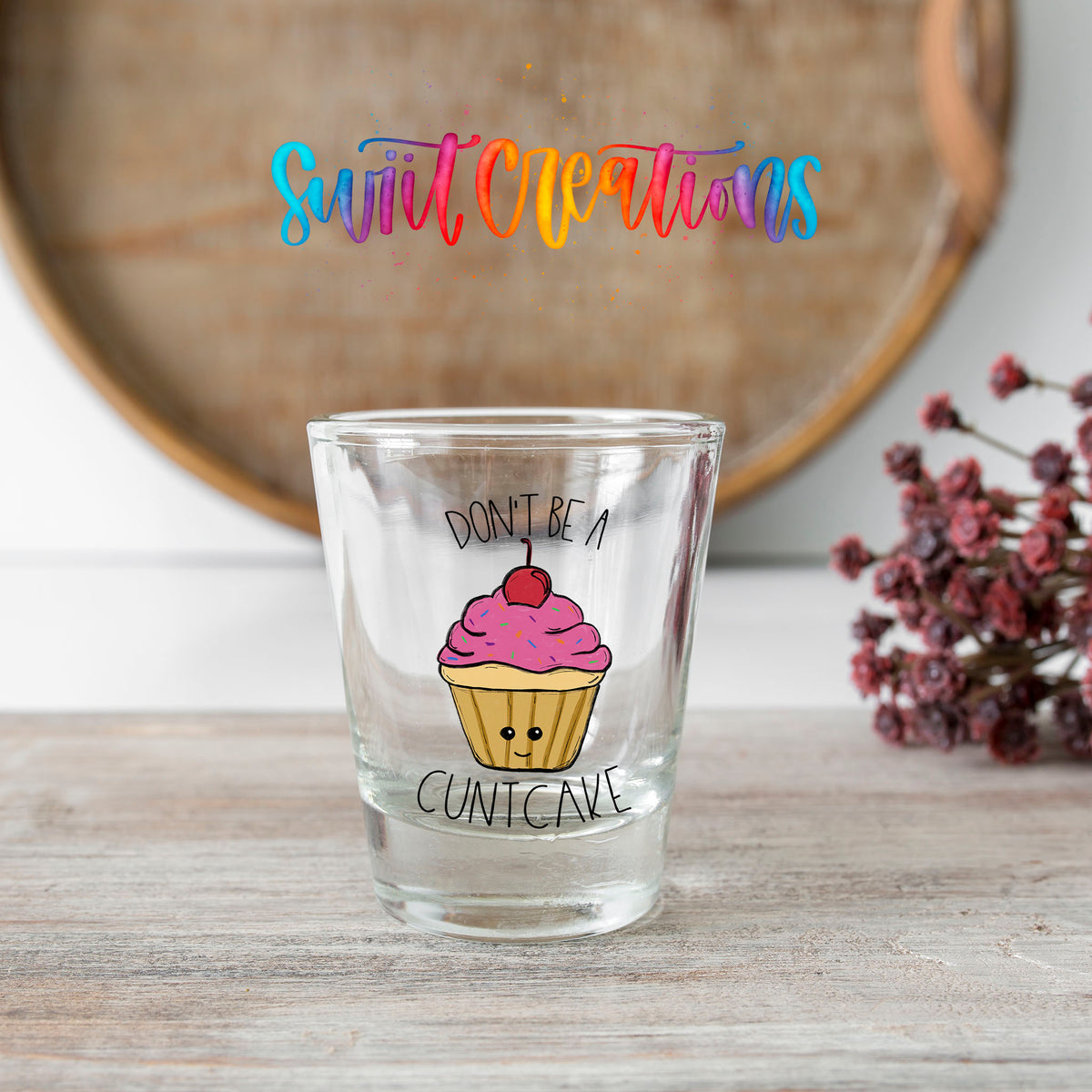 Don't Be a Cuntcake shot glass transfer on a clear glass beside a wooden barrel and dried flowers