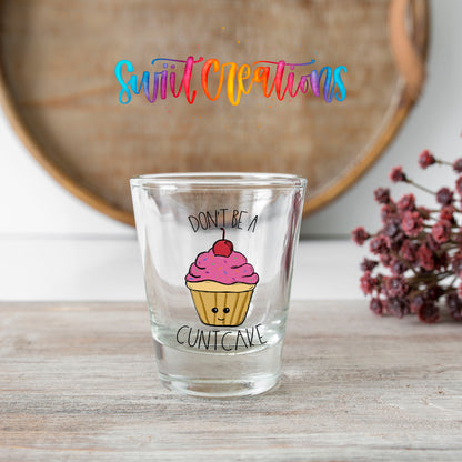 Don't Be a Cuntcake shot glass transfer on a clear glass beside a wooden barrel and dried flowers