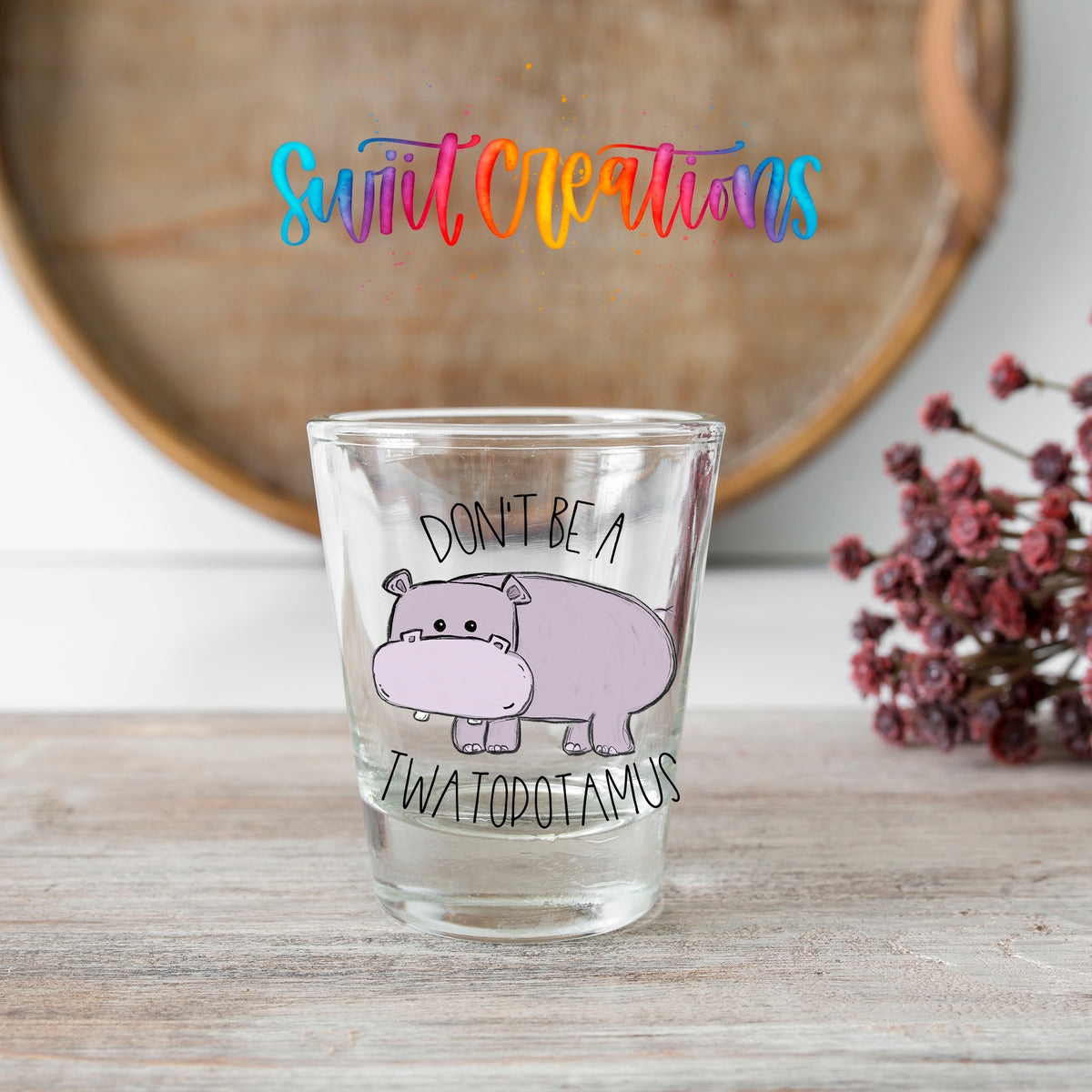 Don't Be a Twatopotamus shot glass transfer on a clear glass beside a wooden barrel and dried flowers