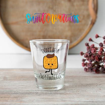Don't Be a Twatwaffle shot glass transfer on a clear glass beside a wooden barrel and dried flowers