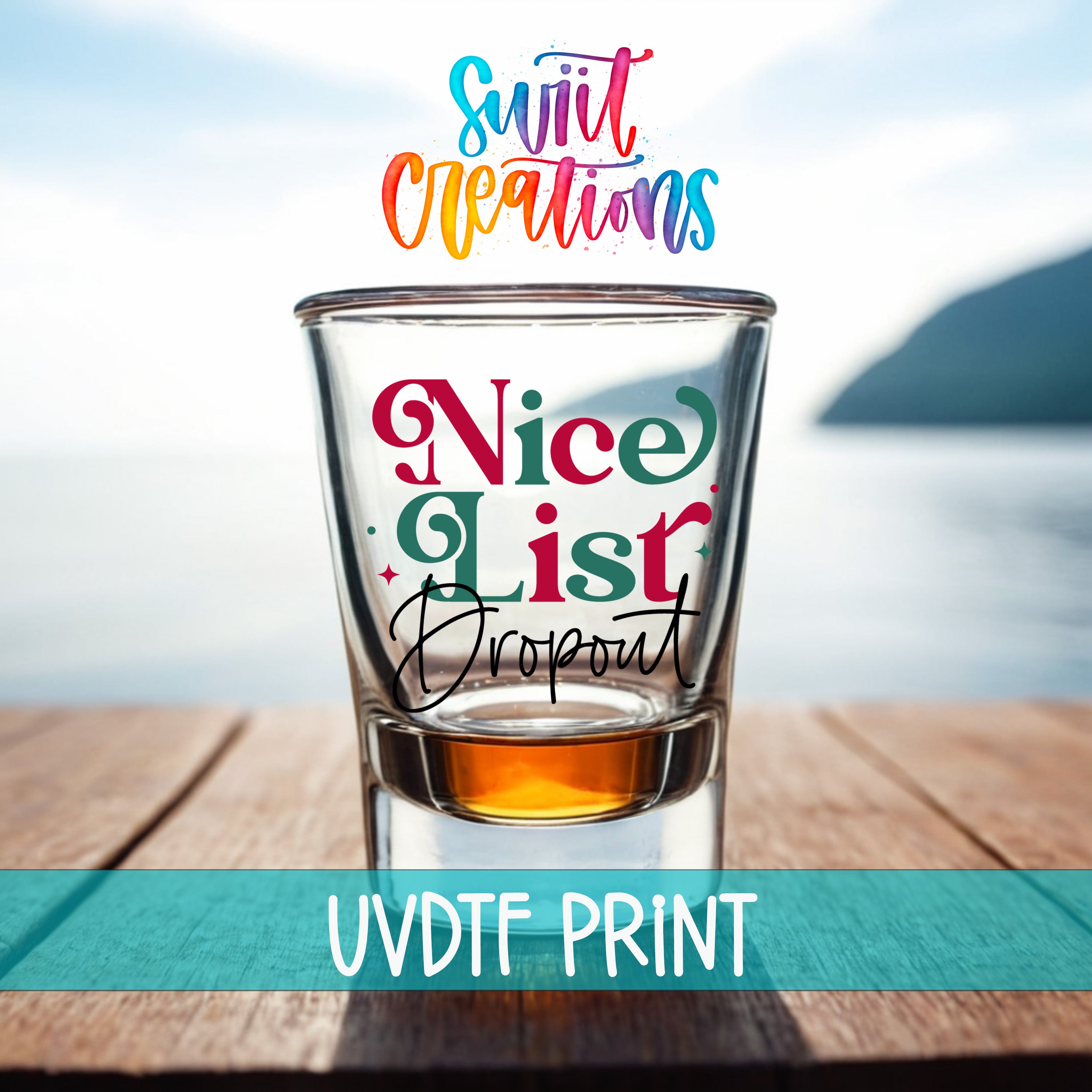a shot glass with a colorful, hand-lettered phrase printed on it, placed on a wooden surface with a blurred background that includes a body of water and a mountain.