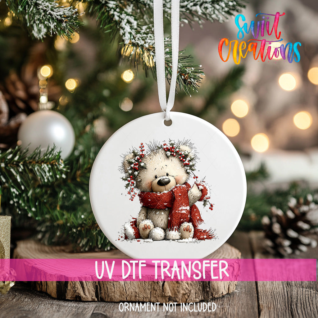 A white ornament with a teddy bear and a red scarf is hanging on a Christmas tree, with a blurred background of pine branches and twinkling lights.
